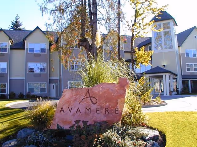 Exterior view of Avamere at St. Helens facility showing a large stone sign with the Avamere logo and name in front of a multi-story building with windows, trees, and landscaped greenery under a clear sky.