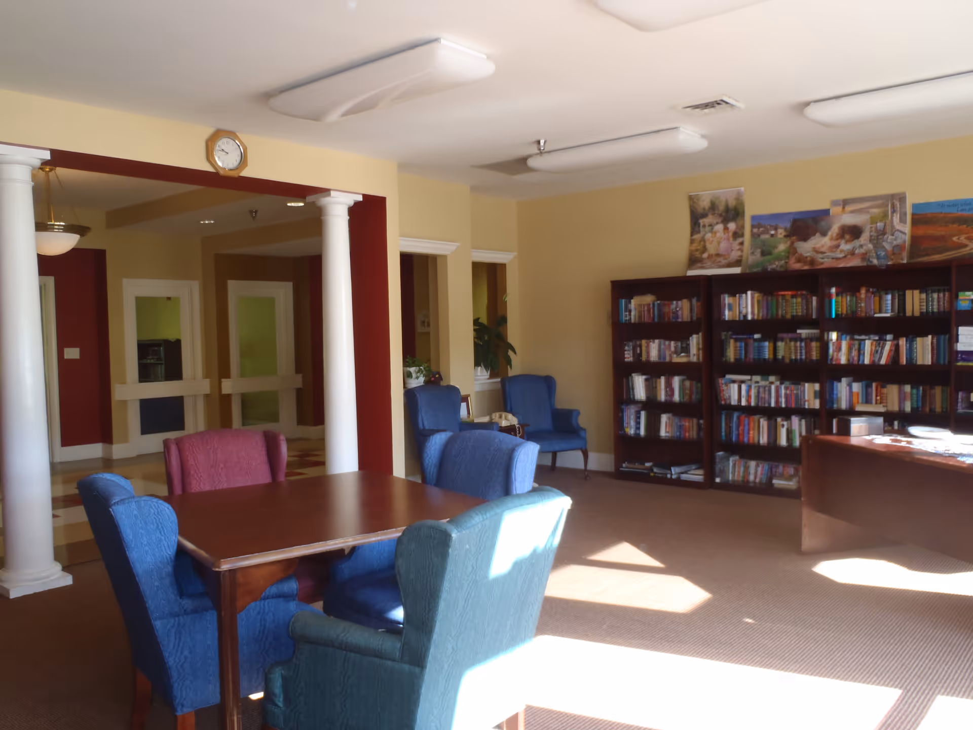 Sunlit common room with a wooden table surrounded by upholstered chairs and bookshelves along the wall.