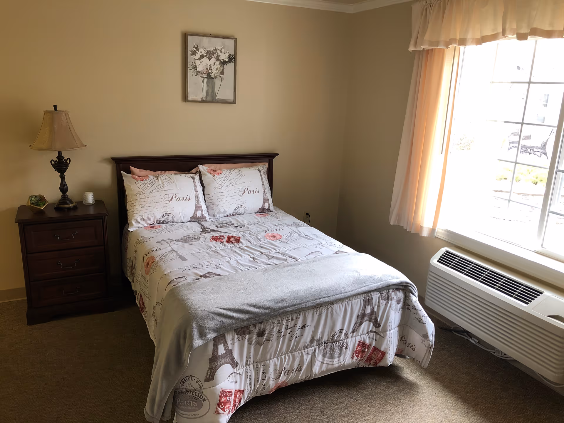 A simple bedroom with a bed covered in Paris-themed bedding, a wooden nightstand with a lamp and small decor items, a framed floral picture on the wall, and a large window with curtains letting in natural light. Below the window is a wall-mounted air conditioning unit.
