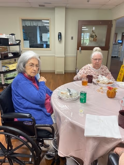 Two elderly women seated at a round table with food and drinks in a nursing home dining area.