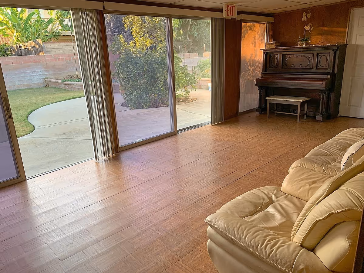 A living room with wooden parquet flooring, a beige leather sofa with a pillow, and an upright piano with a small bench. Large sliding glass doors open to a patio and garden area outside. The room has wood-paneled walls and an exit sign above the door.