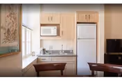 A small kitchen area with light wood cabinets, a white microwave above the countertop, a white refrigerator, a stainless steel sink, and a coffee maker on the counter. Two wooden chairs are partially visible in the foreground, and a window with a white blind is on the left side. A framed picture hangs on the wall near the window.
