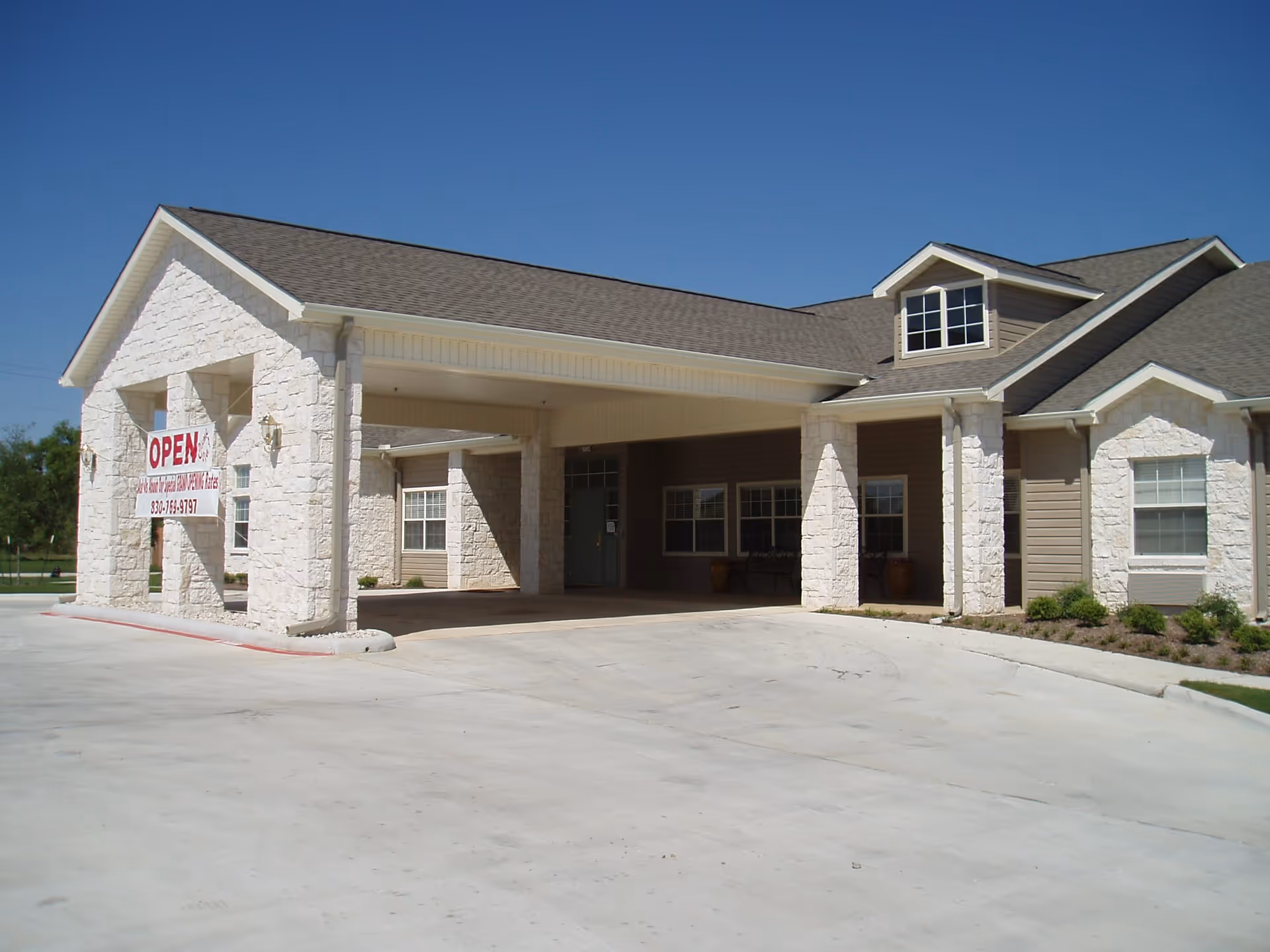 Exterior view of Argent Court Assisted Living facility showing a single-story building with stone and siding facade, a covered entrance with pillars, and a clear blue sky above. There is a sign hanging on the left pillar that says OPEN with a phone number.