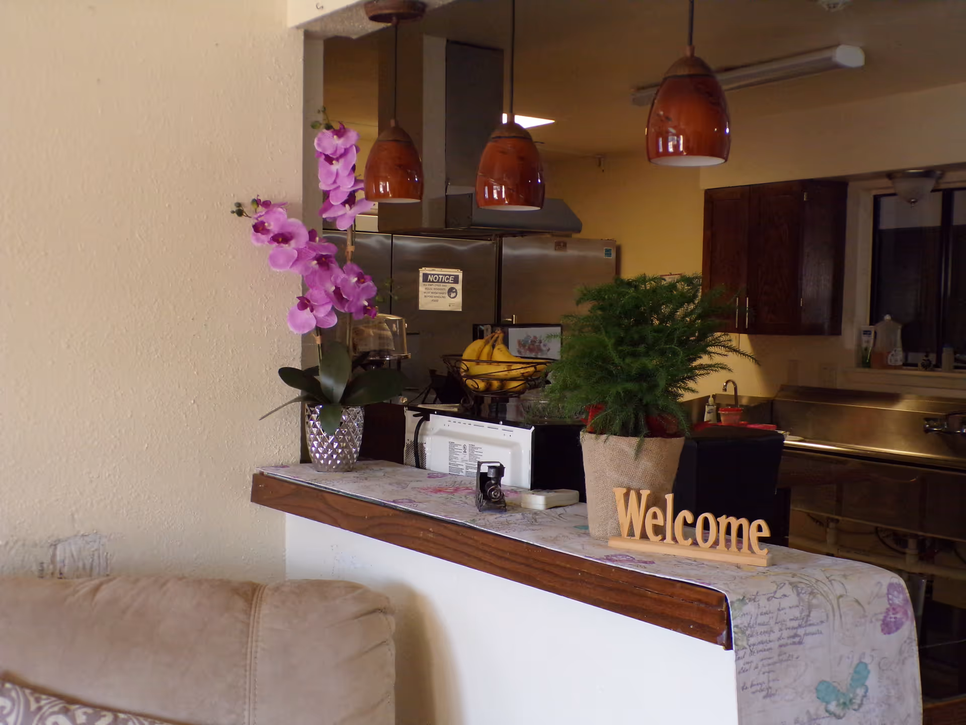 Interior view of a kitchen area with a counter decorated with a purple orchid in a silver vase, a green potted plant in a burlap sack, and a wooden 'Welcome' sign. Behind the counter, there are three hanging brown pendant lights, a stainless steel refrigerator, and wooden cabinets. A beige couch is partially visible in the foreground.