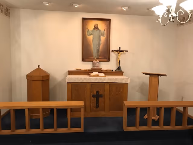 Small chapel-style room with a wooden altar, lectern, pew rails, crucifix, and a large framed image of Jesus on the wall.
