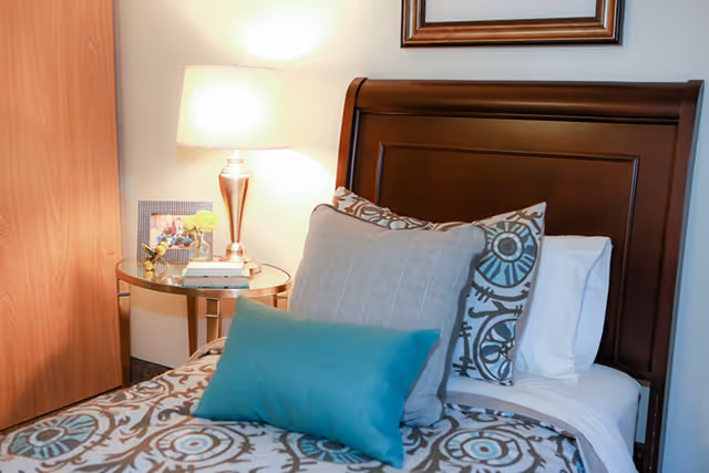 A neatly made bed with patterned bedding and multiple pillows including a teal accent pillow. Next to the bed is a round glass-top side table with a lamp, a framed photo, and a small decorative item. The headboard is wooden with a dark finish.
