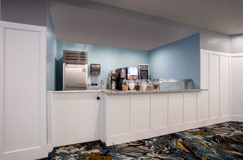 A small serving counter area with a granite countertop, featuring a stainless steel refrigerator, coffee machine, and several jars filled with various snacks. The walls are painted light blue and white, and the floor has a patterned carpet with blue, black, and gold colors.