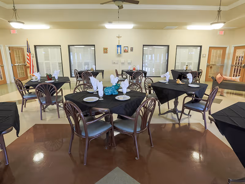 A dining room with multiple round tables covered with black tablecloths, each set with plates, glasses, and white folded napkins. There are wooden chairs around the tables, an American flag in the corner, and a ceiling fan overhead. The room has light-colored walls with windows and doors, and a cushioned bench with pillows along one wall.