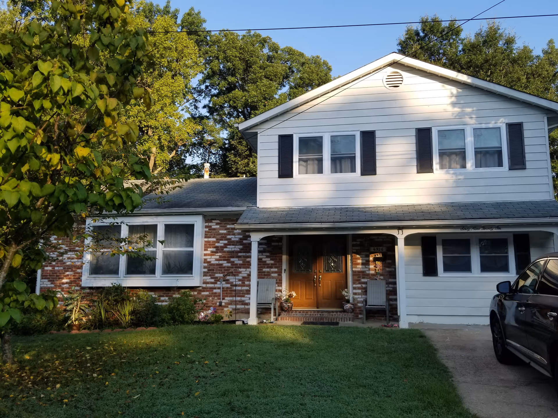 Front exterior view of a two-story house with white siding and brick accents. The house has a covered porch with two chairs and double wooden front doors. There is a green lawn and a tree on the left side, and a black car parked on the driveway to the right.