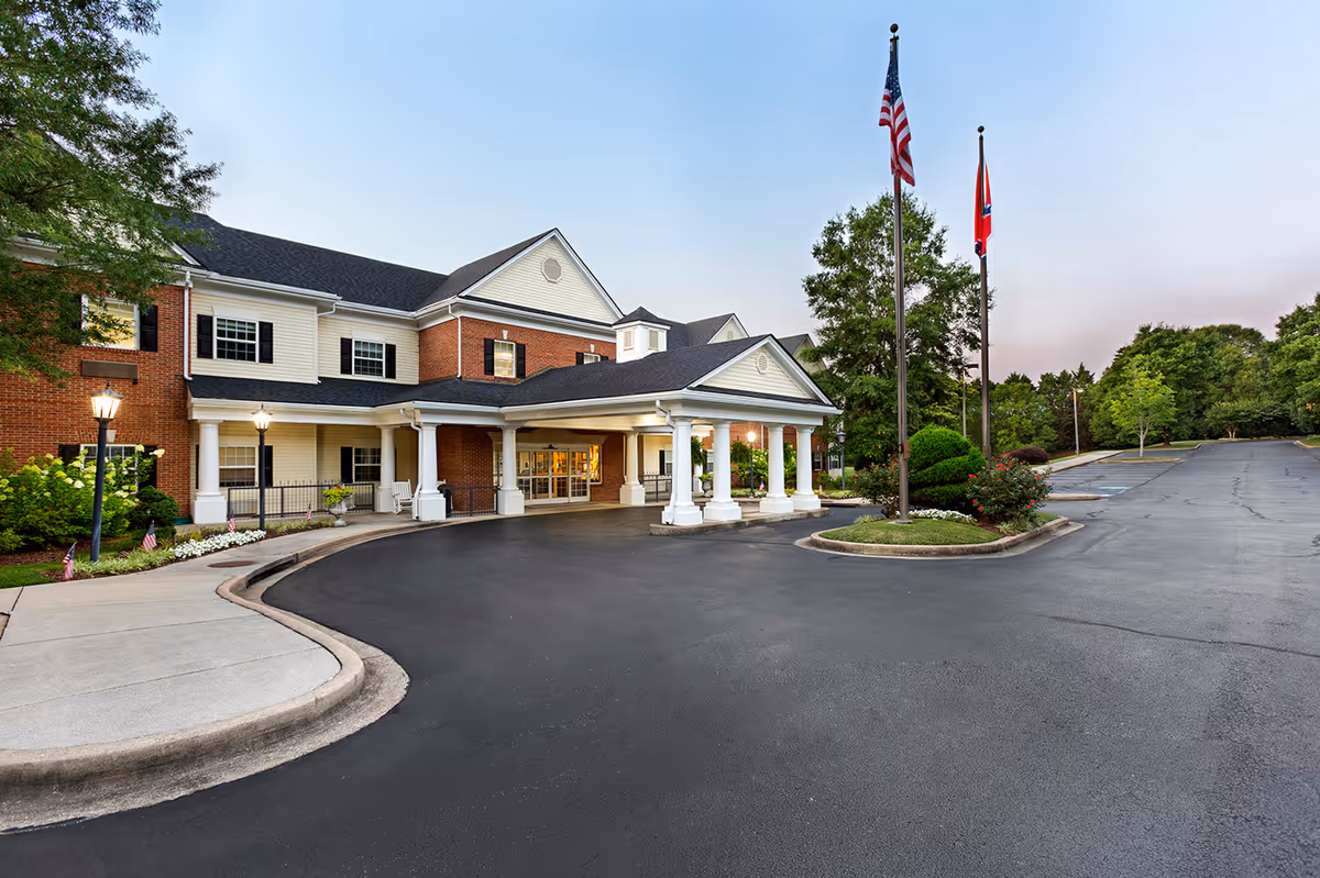 Exterior view of StoryPoint Chattanooga senior living facility showing a large brick and siding building with a covered entrance supported by white columns, two flagpoles with the American and Tennessee state flags, a paved driveway, and surrounding greenery under a clear sky.