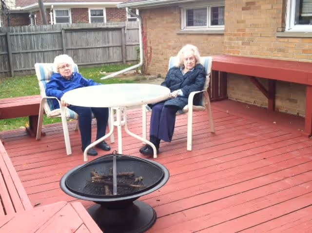 Two elderly women sitting on cushioned chairs around a round white table on a red wooden deck outside. There is a black fire pit in the foreground and a brick building wall with a window and a wooden counter attached to it in the background. A wooden fence and grassy area are visible beyond the deck.