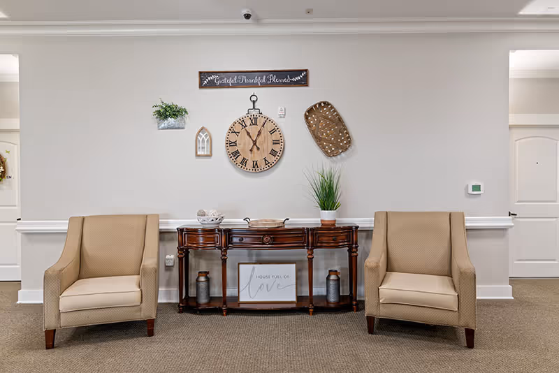 A cozy seating area in a senior living facility with two beige armchairs placed on either side of a dark wooden console table. The table holds decorative items including a plant, a bowl, and a sign that reads 'House full of love.' Above the table on the wall are various decorations including a large round clock, a small plant, a basket, and a sign that says 'Grateful Thankful Blessed.' The walls are light-colored with white trim, and there are doorways visible on both sides of the seating area.