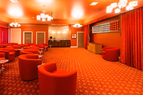 Spacious red-themed communal lounge with rows of red armchairs, chandeliers, and a patterned carpeted floor.