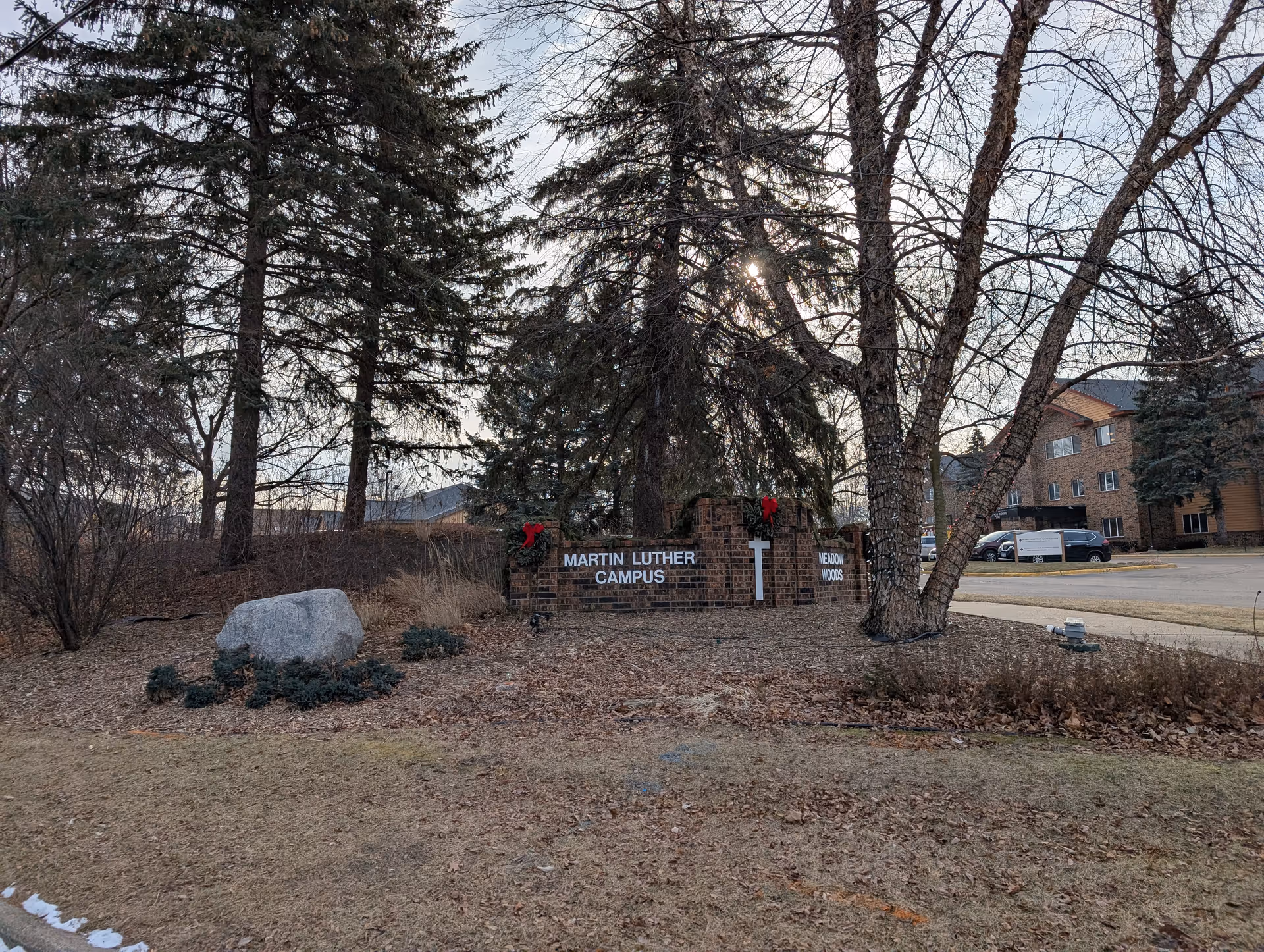 Outdoor view of the entrance sign for Martin Luther Campus, surrounded by trees and landscaping with a large rock and some bushes. There are buildings and parked cars visible in the background.