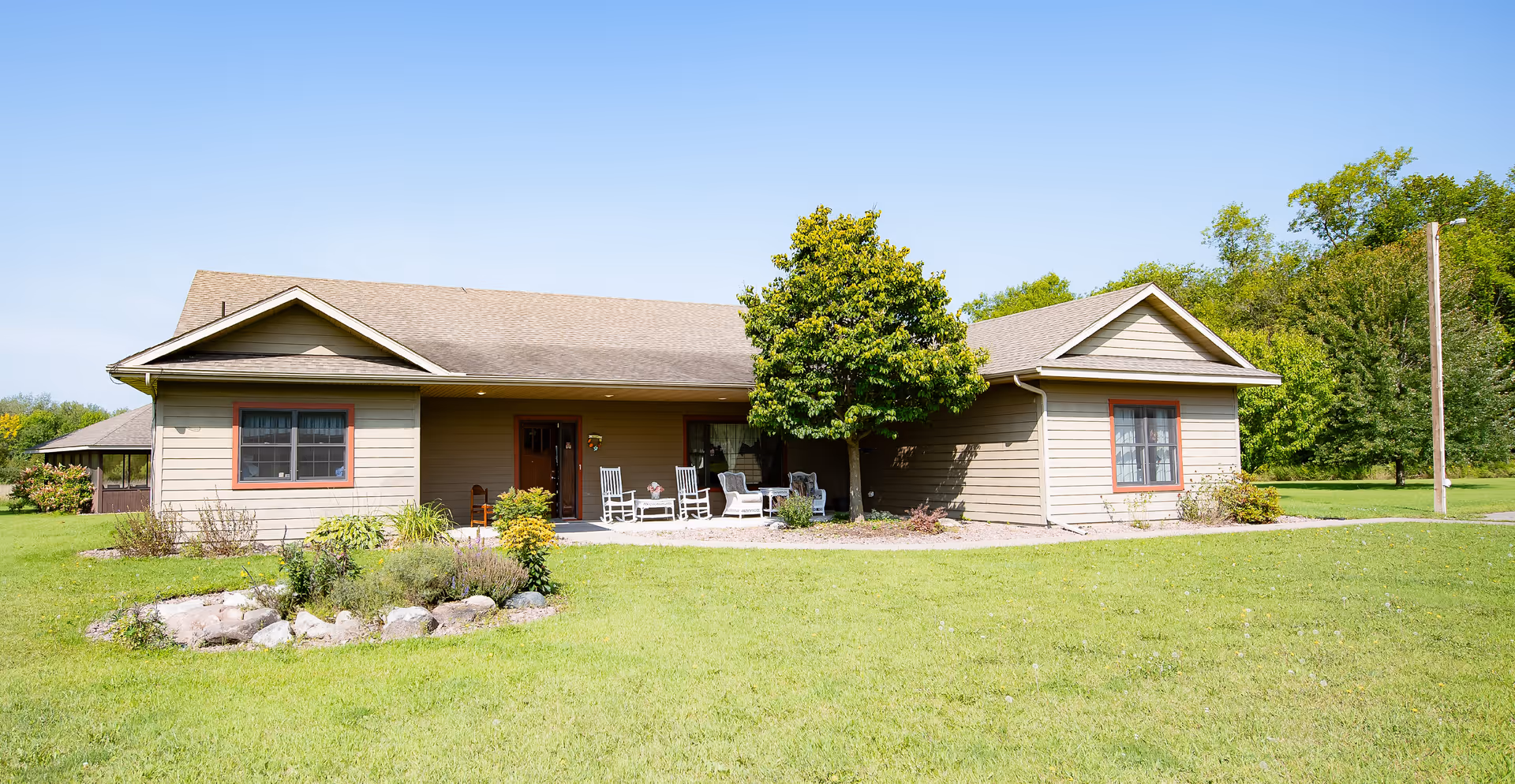 Single-story beige building with a covered front porch, rocking chairs, a tree and landscaped garden set on a large grassy lawn.