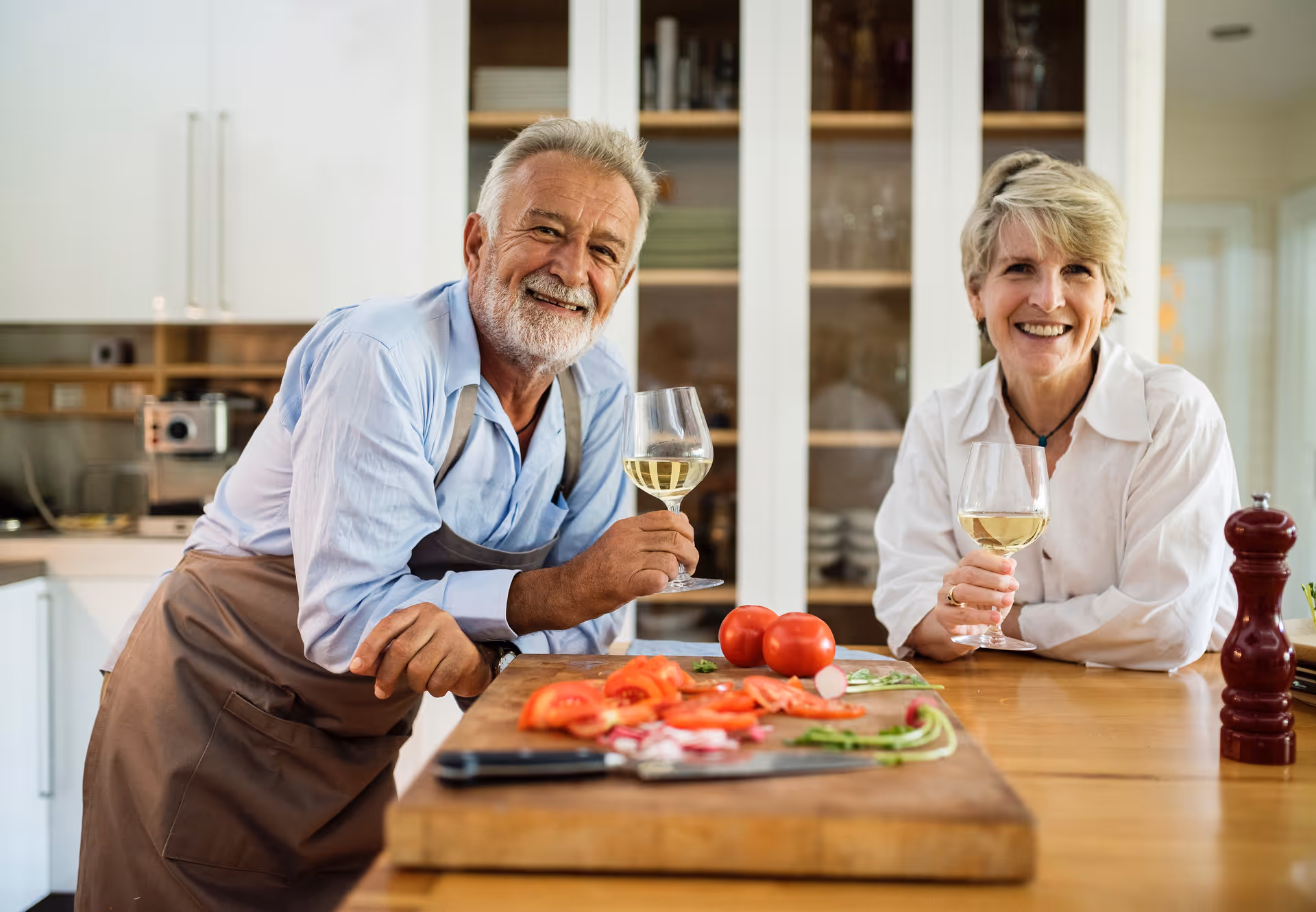 An elderly man and woman smiling and holding glasses of white wine while standing in a kitchen. The man is wearing an apron and leaning on a wooden countertop with sliced tomatoes and other vegetables on a cutting board. The kitchen has white cabinets and shelves with dishes in the background.