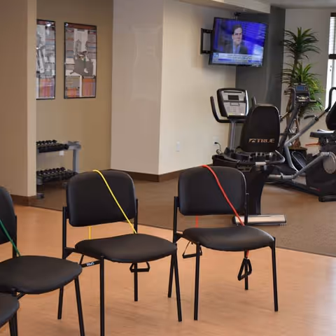 A fitness room with exercise equipment including stationary bikes and dumbbells. Three black chairs with resistance bands attached are arranged in the foreground. A television is mounted on the wall showing a news program. A plant is visible near the window.
