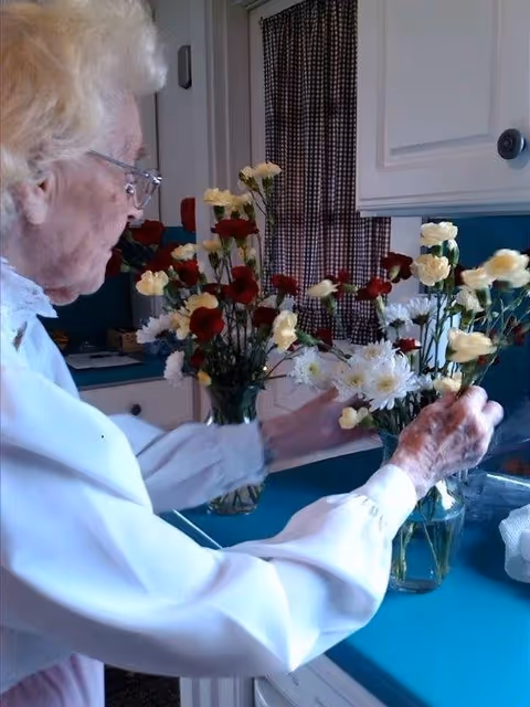 An elderly woman with white hair and glasses arranging red, white, and yellow flowers in glass vases on a blue kitchen countertop. The background shows white cabinets and a window with checkered curtains.