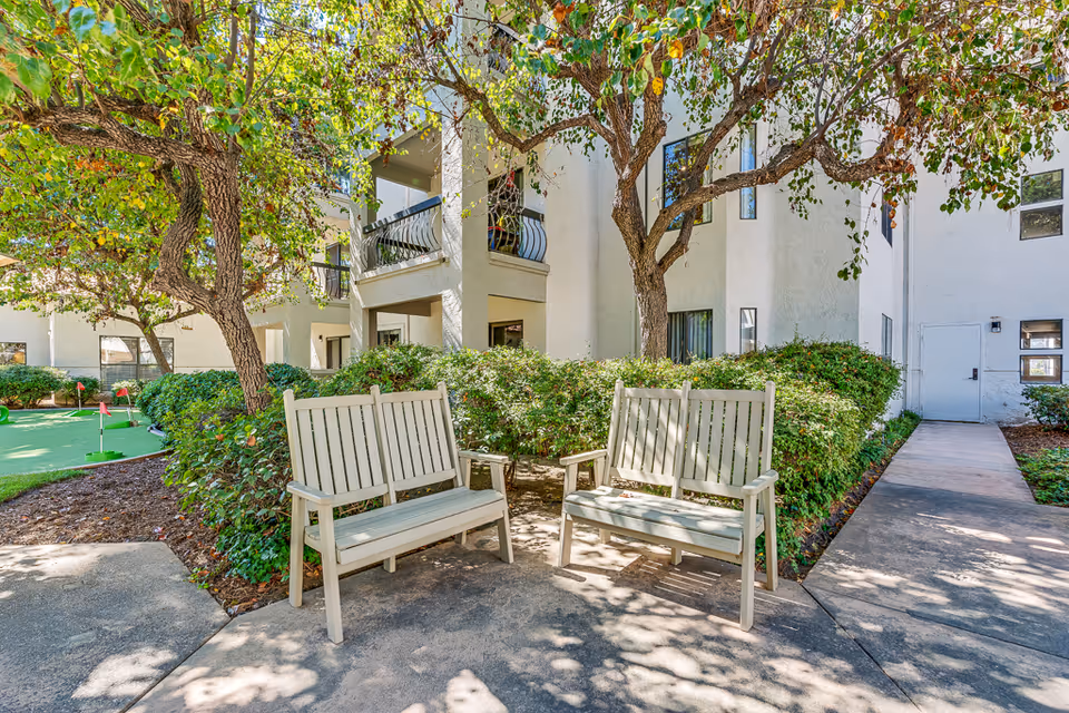 Outdoor seating area in a senior living facility with two wooden benches under leafy trees. The area is surrounded by green bushes and a small putting green with red flags is visible in the background. The building has white walls and balconies.