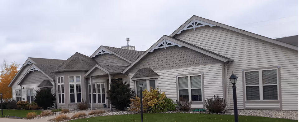 Exterior view of a single-story senior living facility building with beige siding, multiple gabled roofs, and several windows. The building is surrounded by a well-maintained lawn with shrubs and small trees, under a cloudy sky.