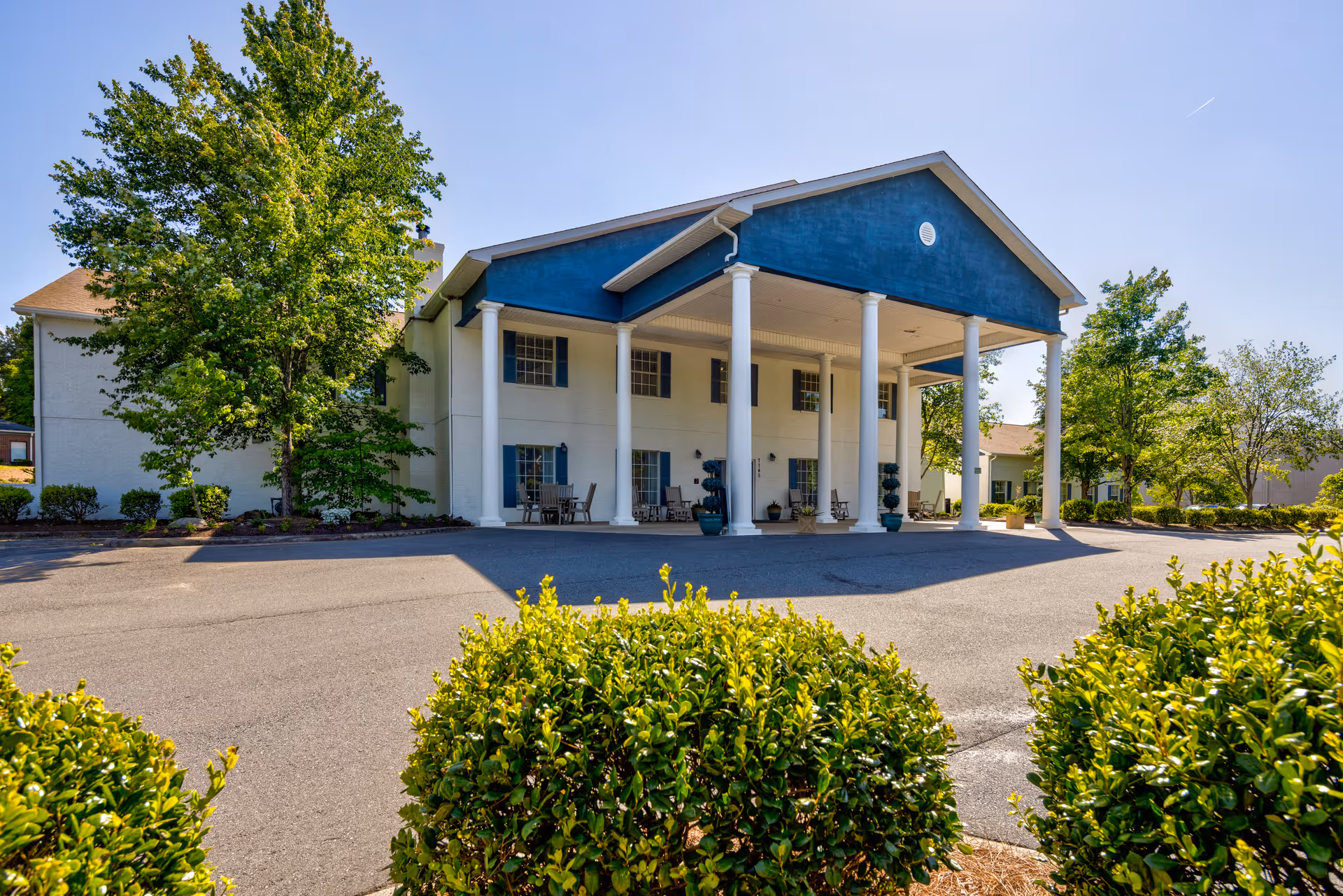 Exterior view of TerraBella Little Avenue facility showing a two-story building with white columns supporting a blue triangular pediment over the entrance. There are several windows with blue shutters, outdoor seating on the porch, and green bushes and trees surrounding the paved driveway in front.