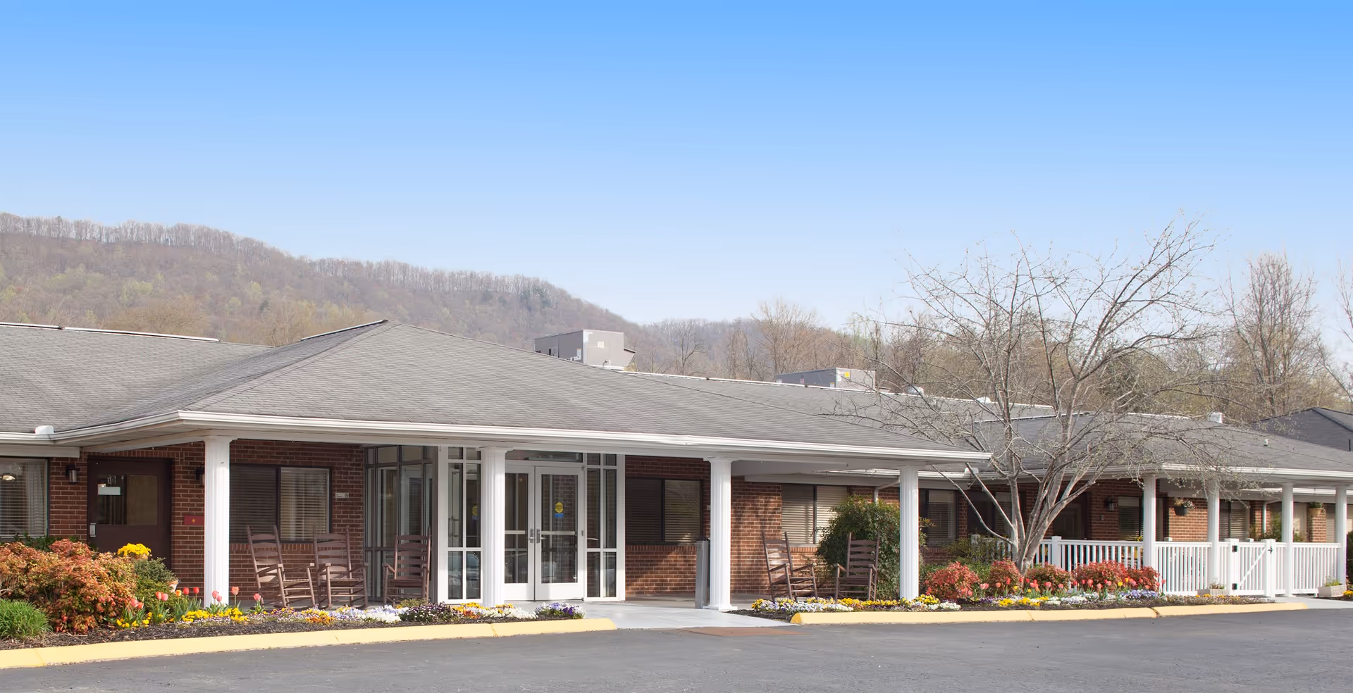 Exterior view of Cumberland Village senior living facility showing a single-story brick building with a covered porch, several rocking chairs, landscaped flower beds, and a backdrop of wooded hills under a clear blue sky.