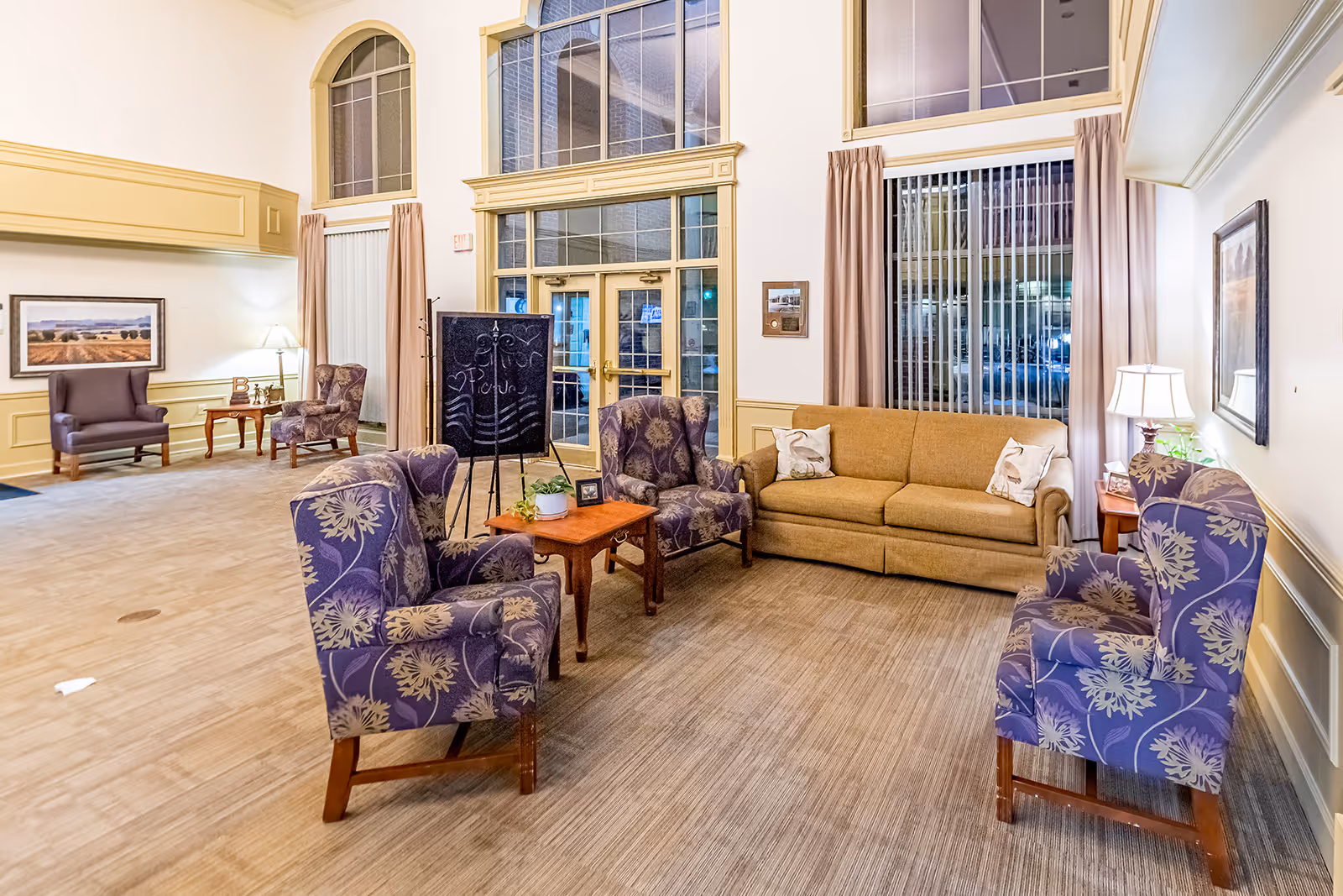 A spacious and well-lit living room area with a beige sofa, four purple floral armchairs arranged around a wooden coffee table, and large windows with vertical blinds and curtains. The room features high ceilings, light-colored walls with decorative molding, a framed landscape picture, and a standing chalkboard with drawings and writing. There are also side tables with lamps and small decorative plants.