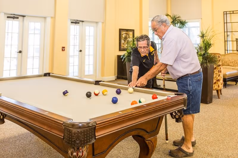 Two elderly men playing pool in a well-lit room with large windows and beige walls. One man is leaning over the pool table aiming a shot while the other watches closely. The room has plants and comfortable seating in the background.
