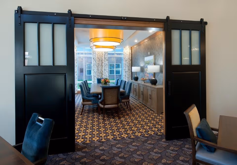 View through black sliding doors into a dining room with a long wooden table surrounded by blue upholstered chairs. The room has patterned carpet, large windows with patterned curtains, two table lamps on a sideboard, and a large circular ceiling light fixture.