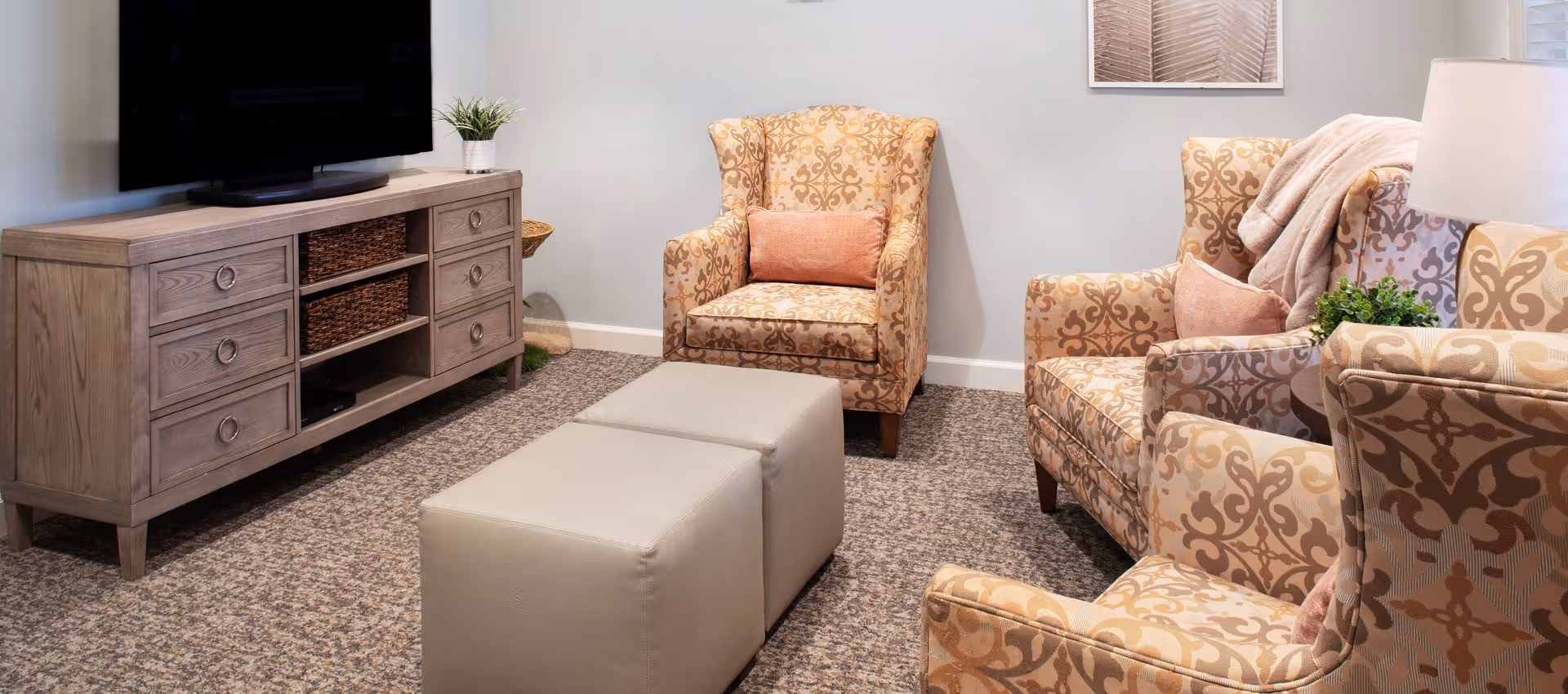Cozy living room with patterned armchairs, a pair of ottomans, and a wooden TV console with a flat-screen television.