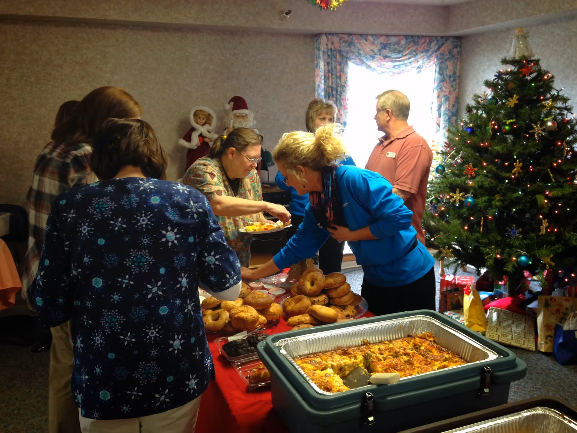 A small group of people serving themselves from a buffet of bagels and hot casserole in a decorated common room with a Christmas tree.