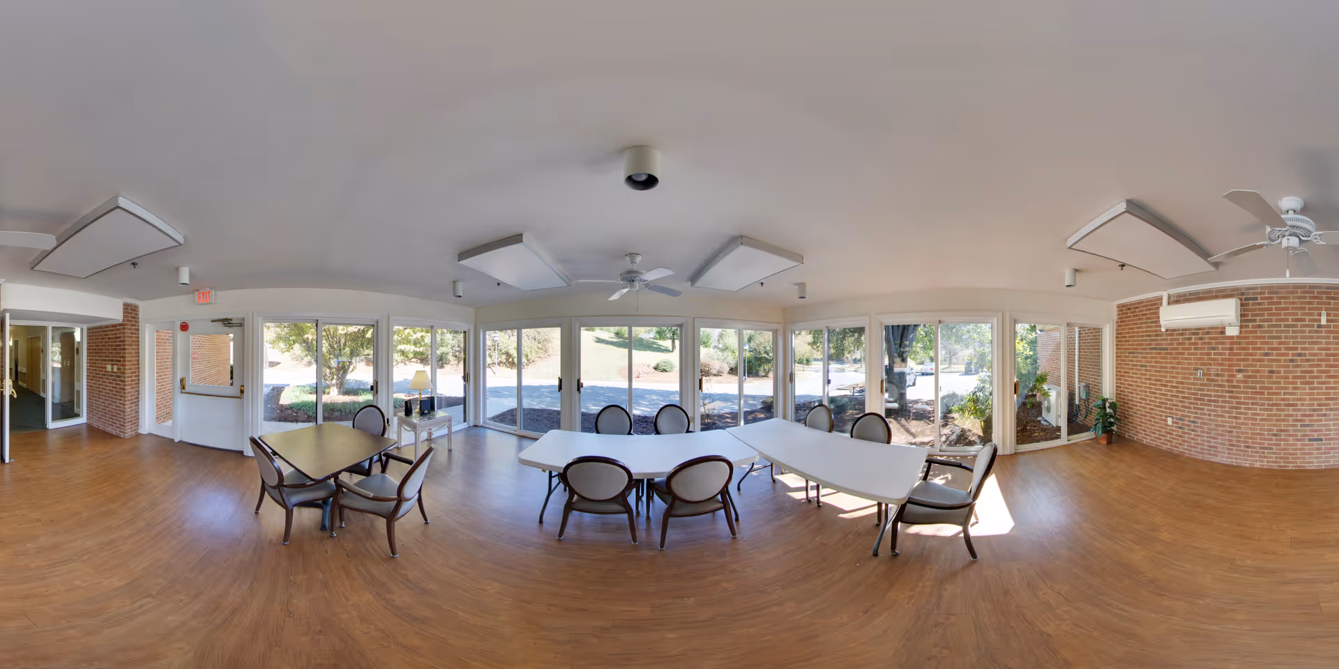 A spacious common area with large windows letting in natural light, several tables with chairs arranged around them, wood flooring, brick walls, ceiling fans, and a view of trees and greenery outside.