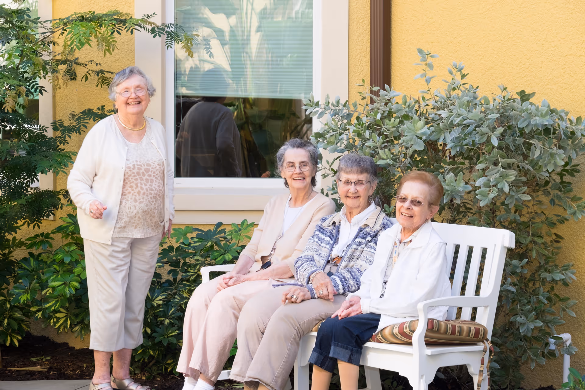Four elderly women smiling outdoors in a garden area. Three women are seated on a white bench with a striped cushion, and one woman is standing beside them. They are in front of a yellow building wall with a window and surrounded by green plants.