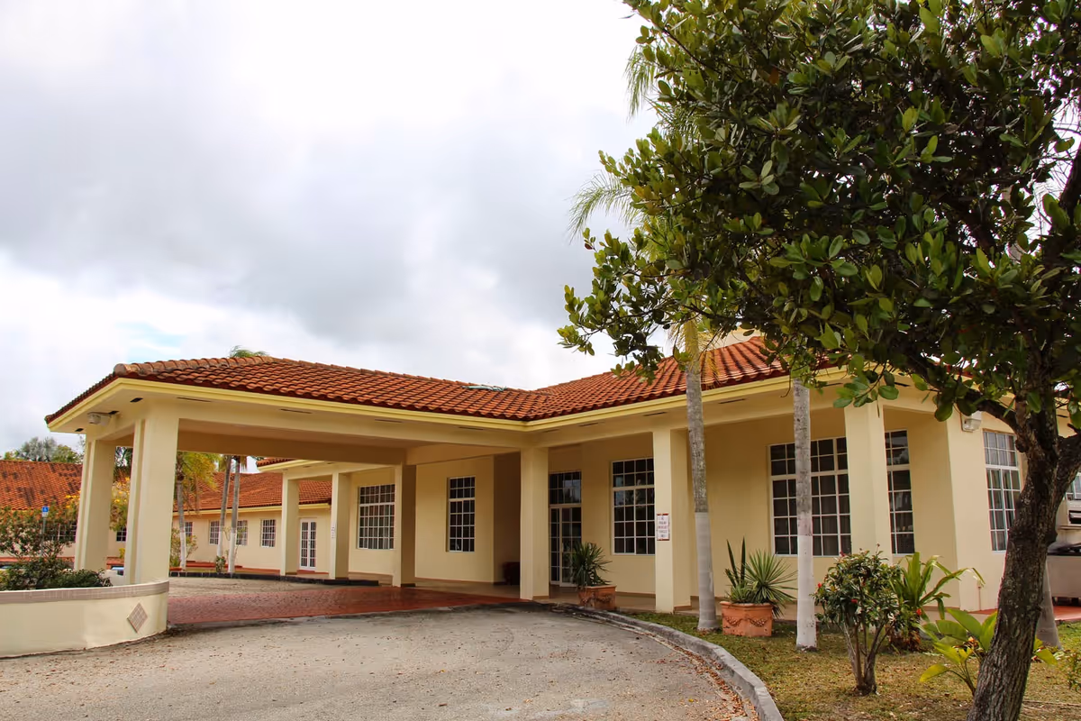 Exterior view of a single-story building with a red tiled roof and beige walls, featuring large windows and a covered driveway entrance. There are trees and plants around the building under a cloudy sky.