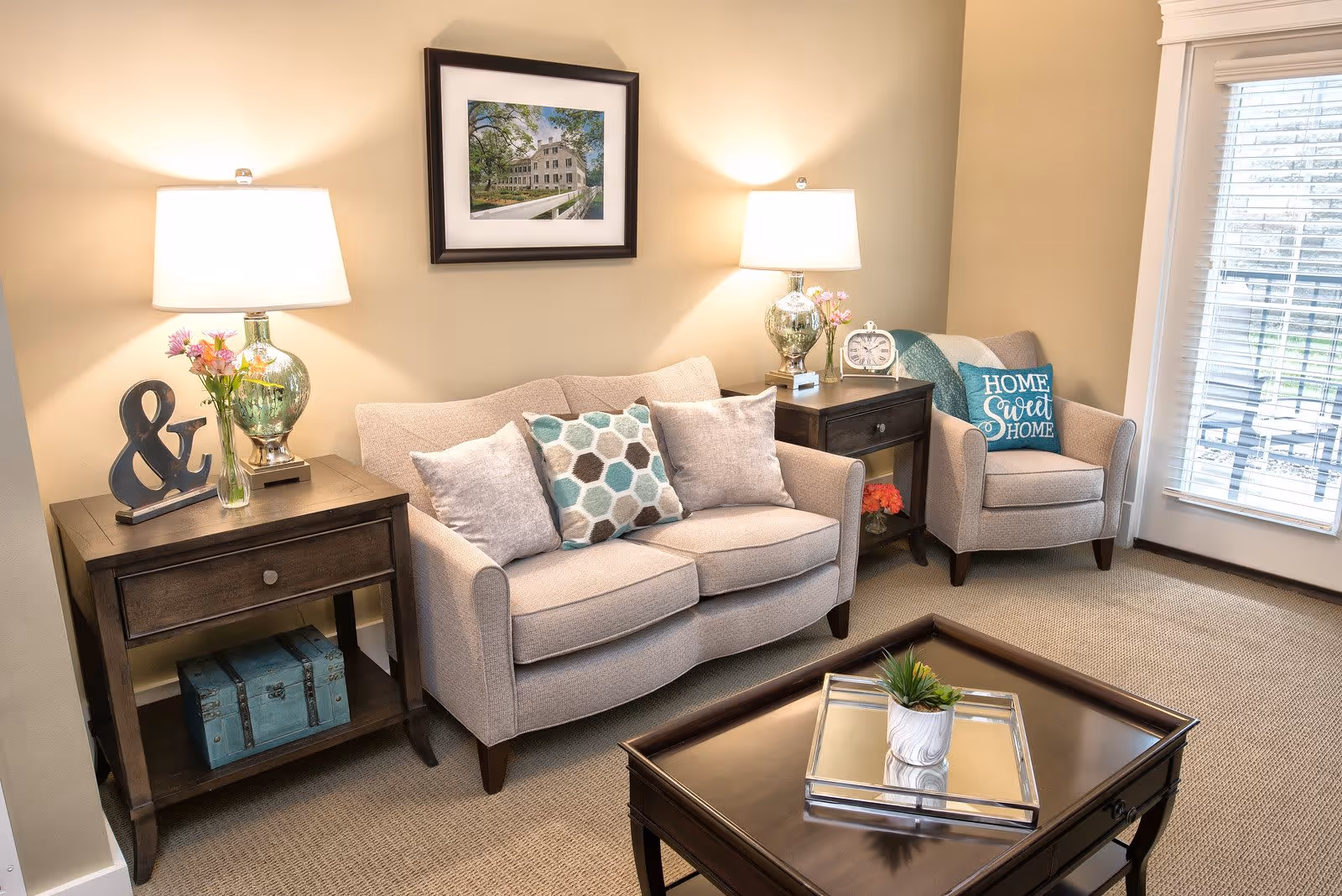 A cozy living room with a beige loveseat and armchair, both adorned with decorative pillows. Two wooden side tables with lamps and flowers flank the loveseat. A framed picture hangs on the wall above the loveseat. A wooden coffee table with a small plant in a white pot sits in front of the seating area. Large windows with blinds allow natural light to fill the room.