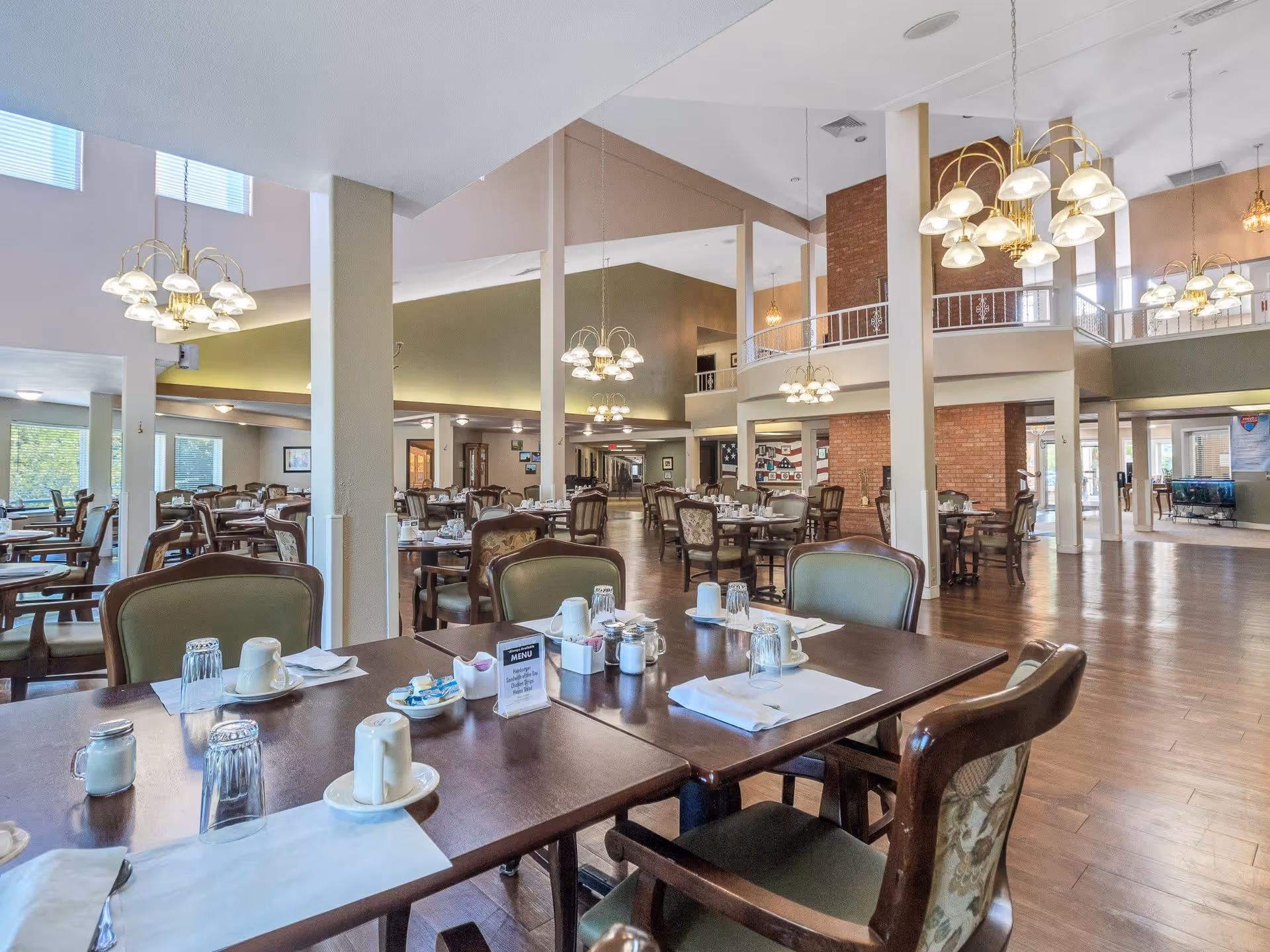 Spacious dining room in a senior living facility with multiple wooden tables and chairs set with cups, glasses, napkins, and condiments. The room features high ceilings, large windows, hanging chandeliers, and a warm, inviting atmosphere with wooden flooring and a brick accent wall.