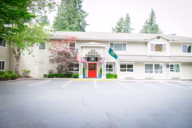 Exterior front view of a two-story senior living facility building with beige siding, a red entrance door under a small portico, American and Washington state flags, and surrounding trees and plants. The parking lot in front is empty.