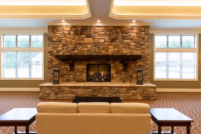 Cozy lounge with a stone fireplace centered between two large windows, a beige sofa facing it and side tables.