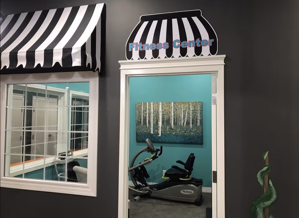 View of a fitness center room through an open doorway with a black and white striped awning above the door and window. Inside the fitness center, there is a NuStep exercise machine and a painting of birch trees on a teal wall.