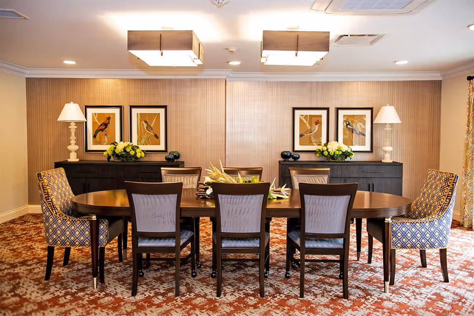 A formal dining room with a large oval wooden table surrounded by eight chairs, two of which have patterned upholstery. The room features a patterned carpet, two black sideboards with white lamps and floral arrangements, and framed bird artwork on the wall.