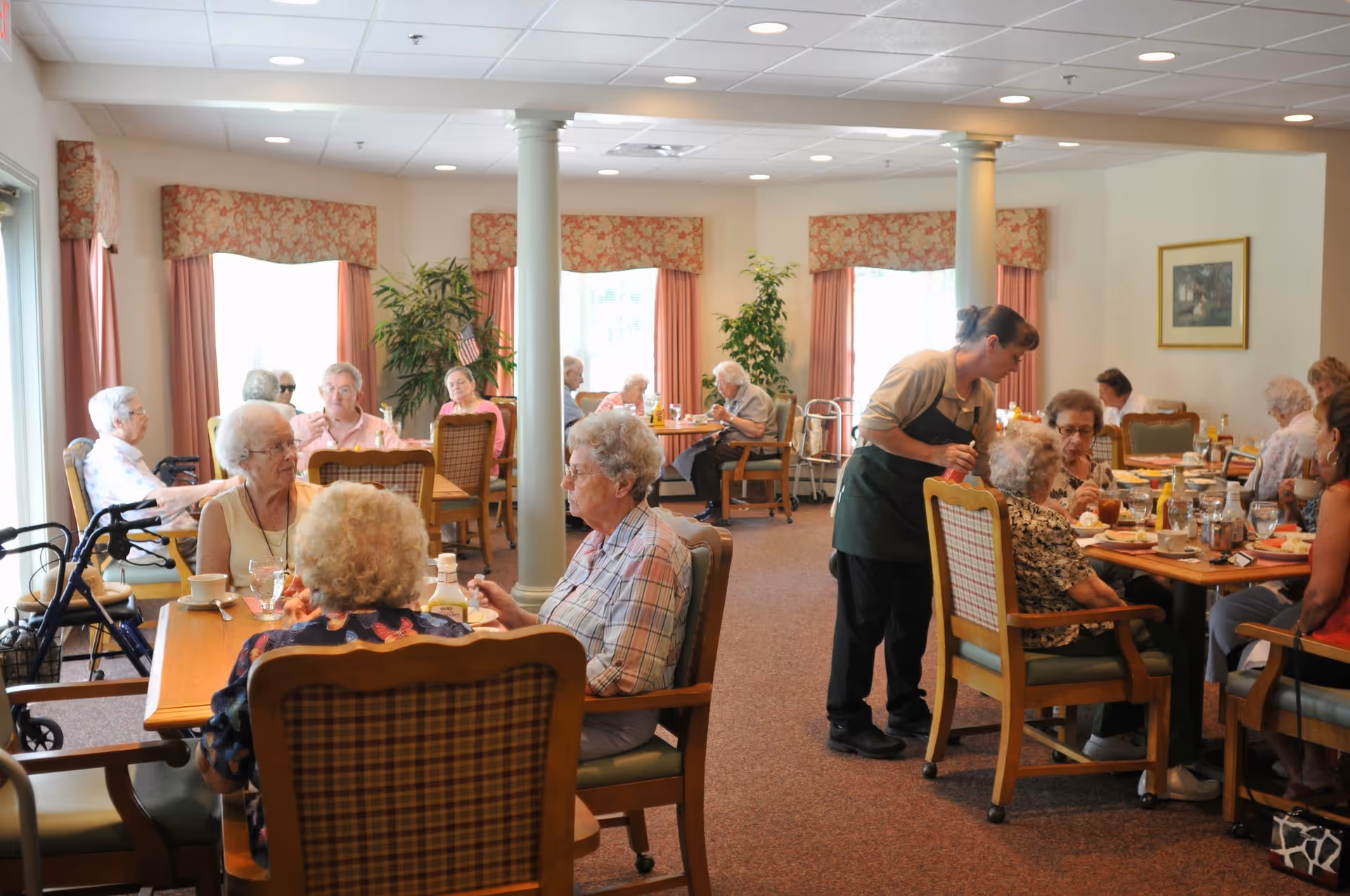 A dining room in a senior living community with elderly residents seated at tables, eating and conversing. A staff member is serving food to one of the tables. The room is well-lit with large windows covered by floral curtains, and there are plants and framed artwork on the walls.