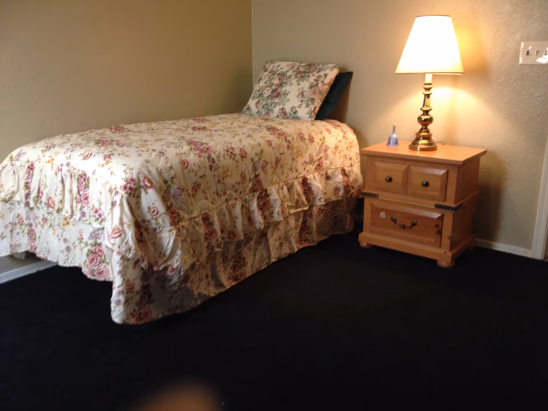 Single bed with a floral bedspread and a wooden nightstand with a lamp in a simple bedroom.