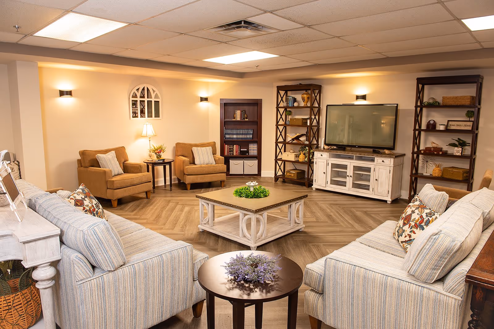 A cozy living room with two striped sofas facing each other, a square wooden coffee table with a green centerpiece, two brown armchairs with pillows, a small round side table with a lamp and flowers, two tall wooden bookshelves filled with decorative items and books, and a white TV stand with a flat-screen television mounted above it. The room has warm lighting and a wood-patterned floor.