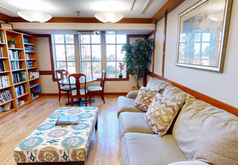 Sunny common lounge with a sofa, patterned ottoman, bookshelves, and a small table and chairs by large windows.