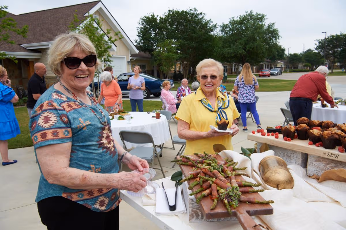 Two elderly women smiling and standing near a table with appetizers and muffins at an outdoor gathering in a senior living community. Other people are socializing and sitting at round tables with white tablecloths in the background, with houses and trees visible.