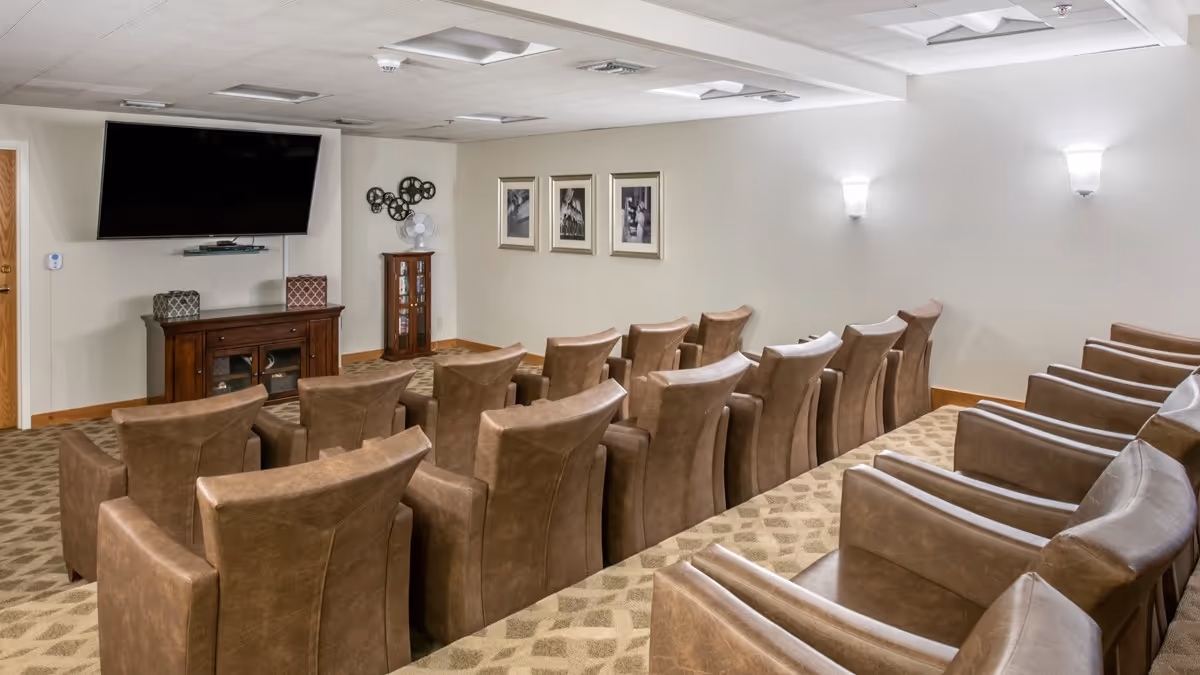 A small theater room with three rows of brown leather chairs facing a large flat-screen TV mounted on the wall. The room has beige walls, carpeted floor with a patterned design, wall sconces for lighting, and framed black and white pictures on the wall.