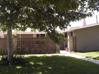 Outdoor view of a residential facility with a tree in the foreground, a grassy lawn, a walkway, and a building with a brick lattice wall and an American flag on the roof.