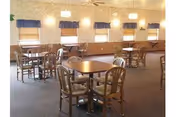 A dining room with multiple wooden tables and chairs arranged neatly. The room has several windows with blue valances and beige blinds, and the walls are decorated with light sconces. The floor is carpeted, and the overall atmosphere is warm and inviting.