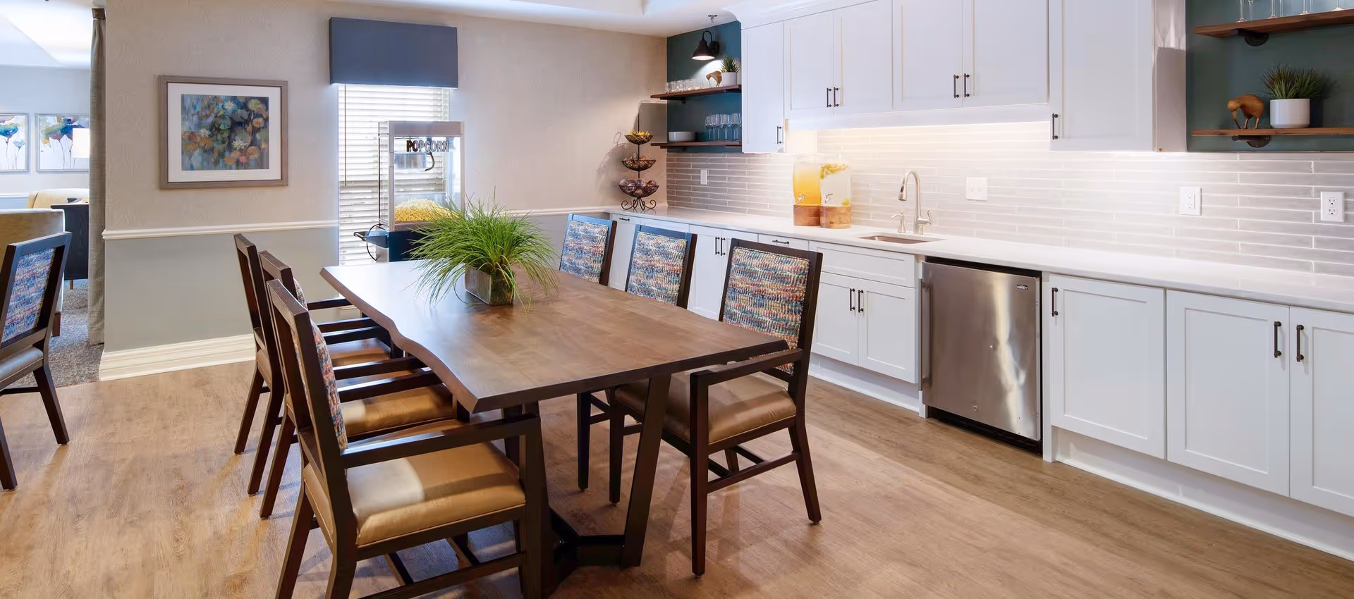 Communal dining area with a long wooden table and chairs beside a kitchenette with white cabinets and a stainless dishwasher.
