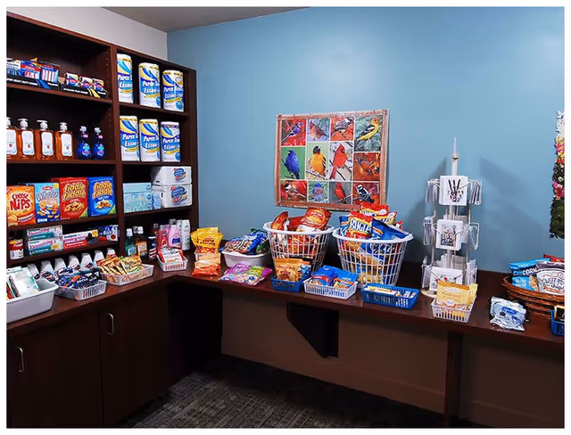 A small snack shop area inside a facility with shelves and baskets filled with various snacks such as chips, crackers, and paper products. The walls are painted light blue and there is a colorful bird-themed quilt hanging on the wall. A rotating rack with greeting cards is also visible on the right side.